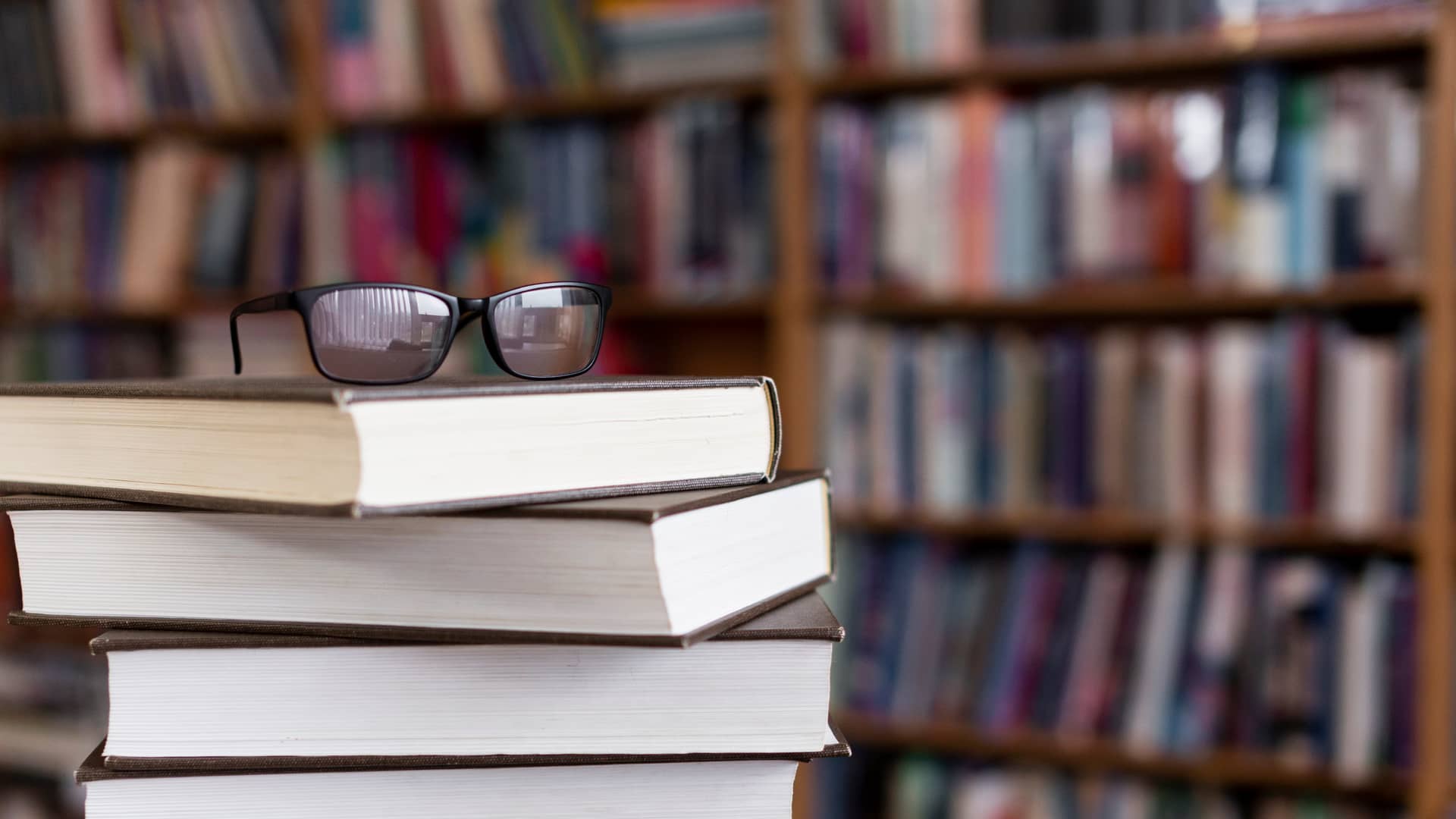 Torre de libros con unas gafas encima, y una librería llena de tomos para la formación financiera de fondo