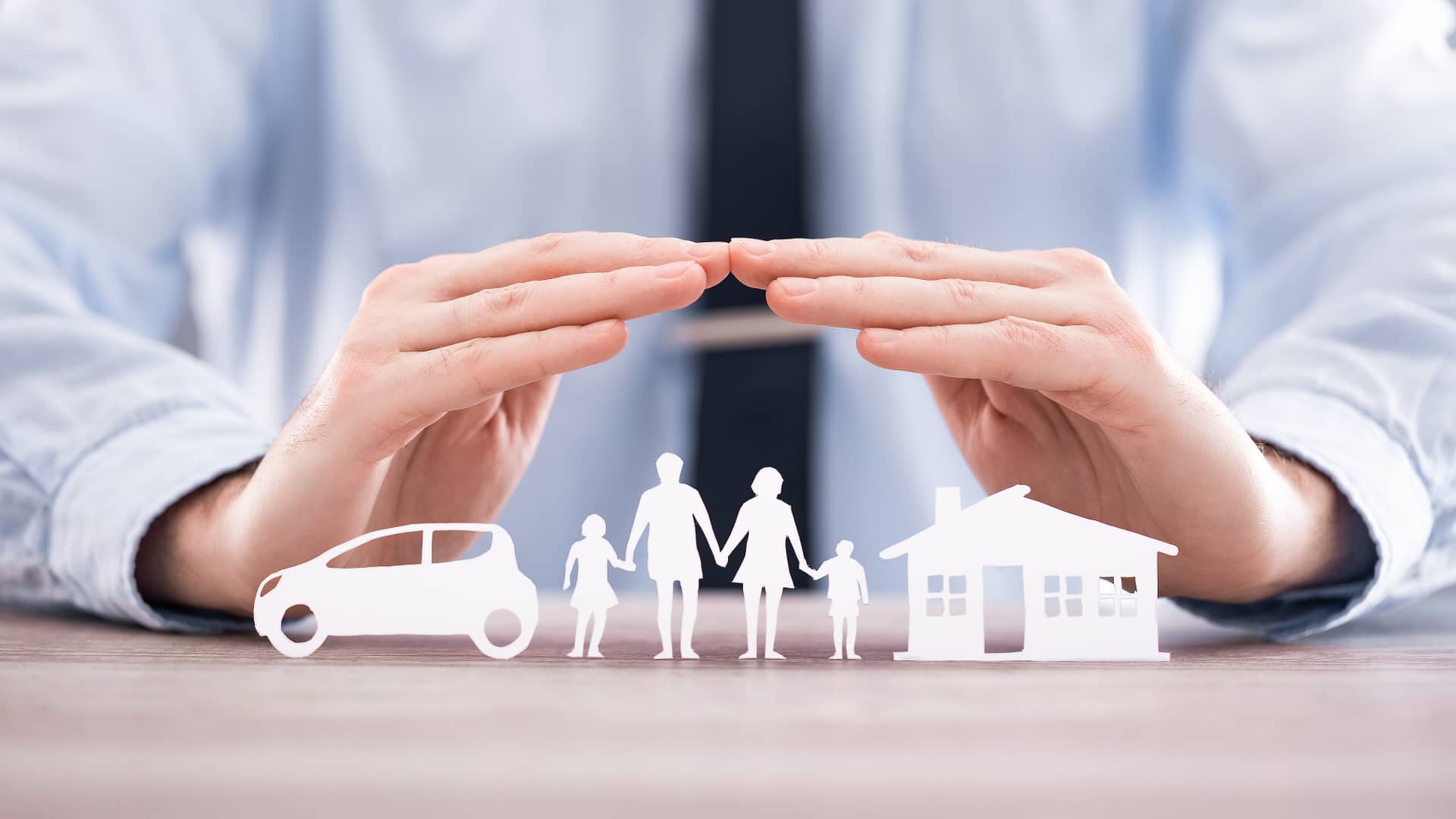 Persona protege con sus manos las siluetas de papel de una familia su hogar y su coche, representando los seguros vinculados a hipoteca