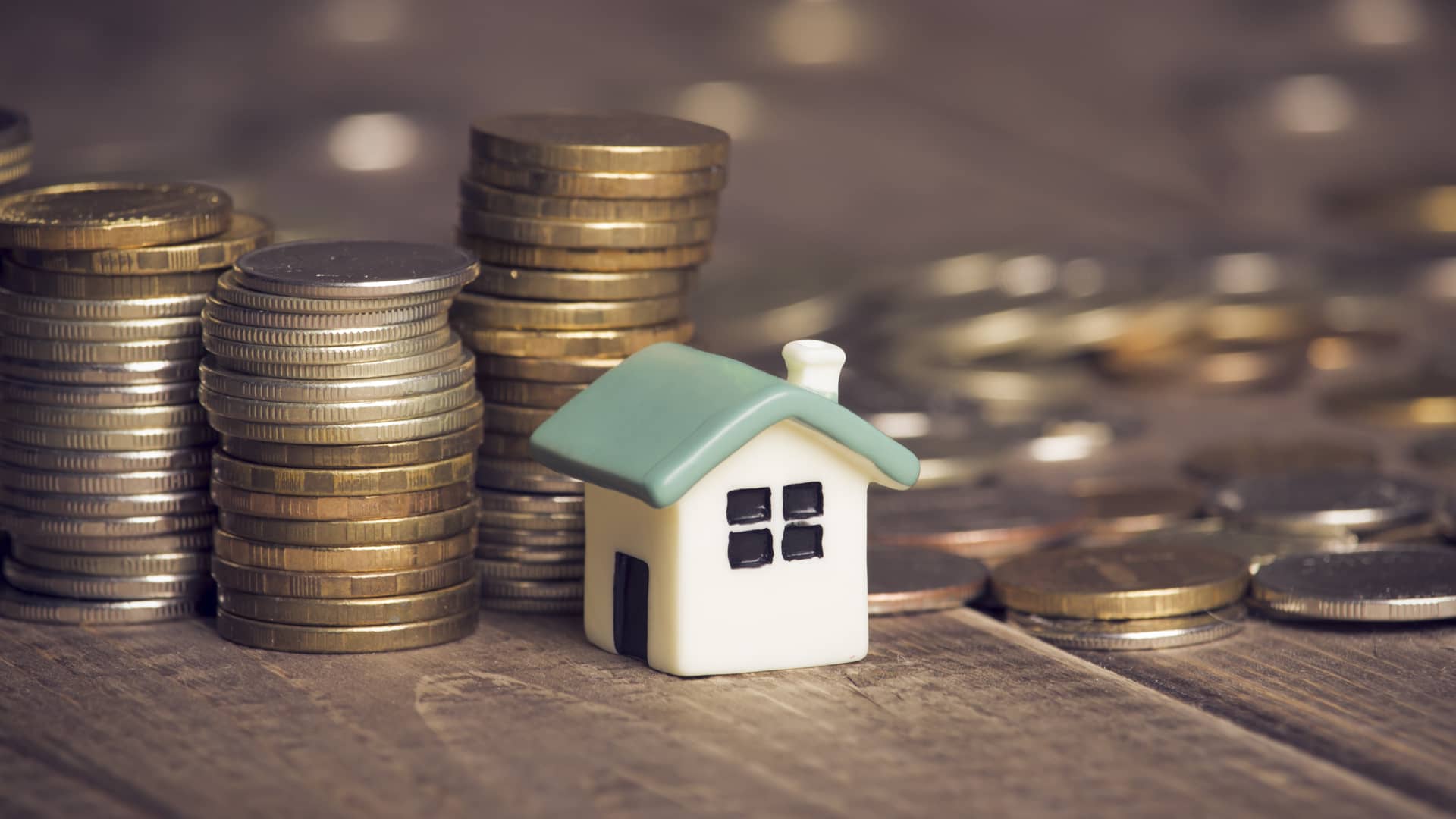 Montañas de monedas rodeando una pequeña casa de juguete simulando cuando se dejan de pagar los intereses de una hipoteca