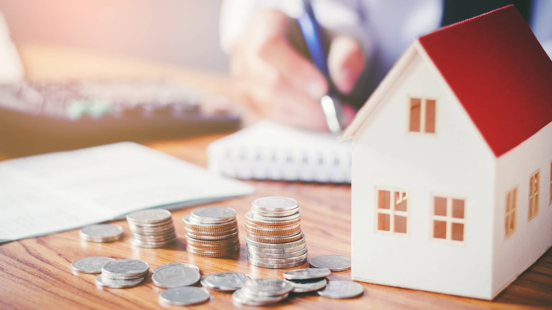 Persona haciendo cuentas  junto a una casita de juguete y dinero símboliza el estudio previo que realiza el banco antes de conceder la hipoteca